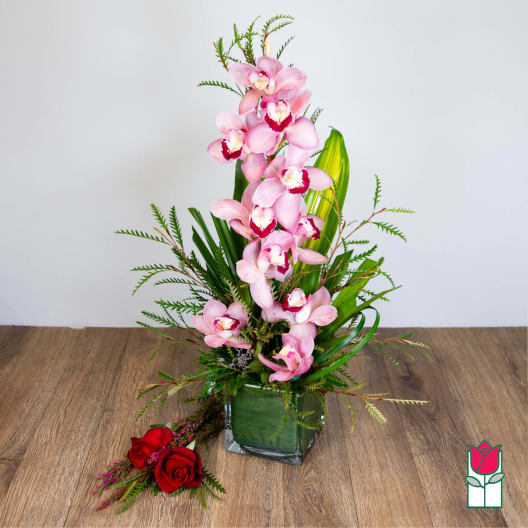 Tall arrangement of pink cymbidium orchids in a green-lined glass cube with two red roses beside it