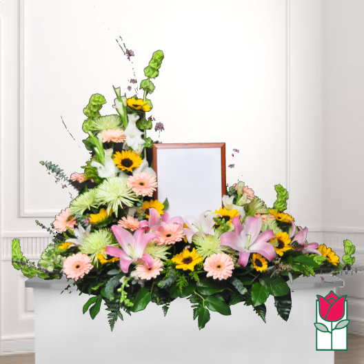 Symmetrical floral funeral spray with pink lilies, daisies, and sunflowers around a framed center