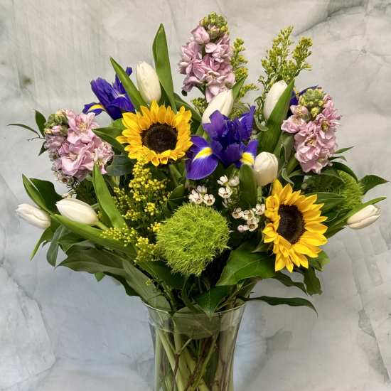 Mixed bouquet with sunflowers, purple irises, white tulips, and pink blooms in a glass vase