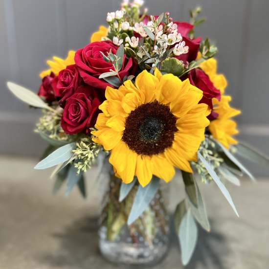 Arrangement of yellow sunflower, red roses, and small white blooms in a clear glass jar vase