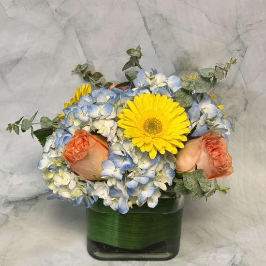 Bouquet of blue and white hydrangeas with yellow gerbera daisies and peach roses in a glass vase