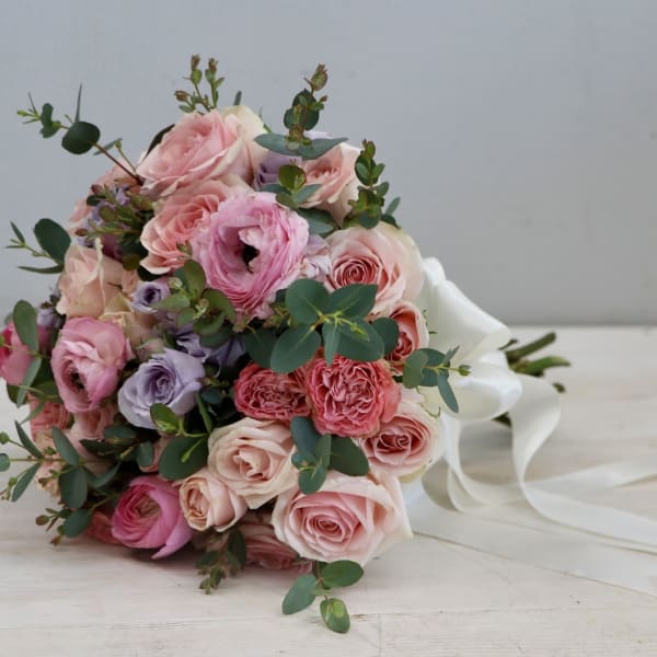 Pastel pink and peach rose bouquet with lavender blooms and white ribbon trailing on a light surface