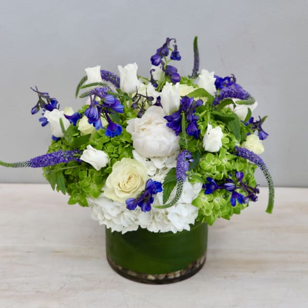 Low round arrangement of white and blue flowers with green accents in a glass vase wrapped with a green leaf.