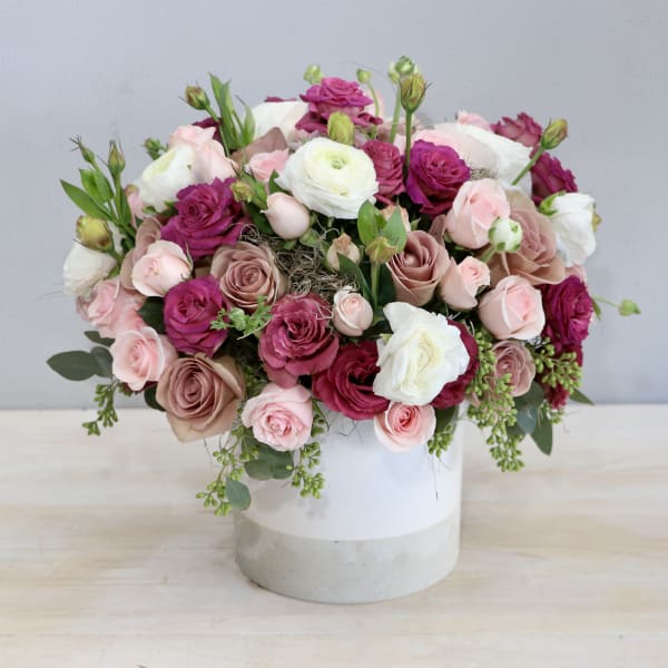 Low arrangement of pink, mauve, and white roses and ranunculus in a white cylinder vase