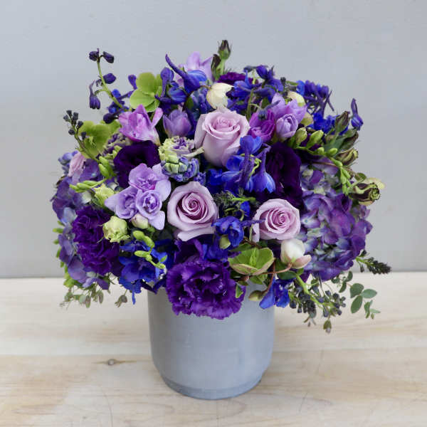 Round arrangement of purple, blue, and lavender flowers in a gray cylinder vase