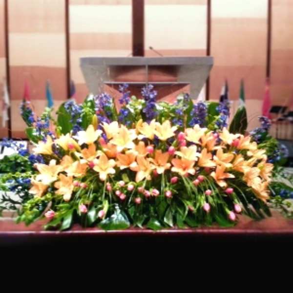 Low altar spray of peach lilies, blue flowers, and pink buds in a lush horizontal display.