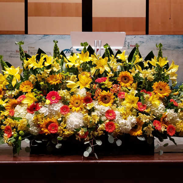 Large stage arrangement of yellow lilies, orange gerbera daisies, and white hydrangeas in a low black container