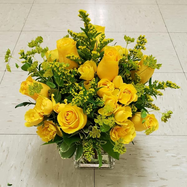 Yellow roses arranged in a clear square vase with green filler flowers