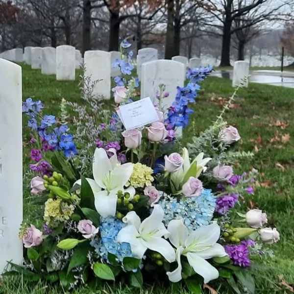 Sympathy arrangement of white lilies, pastel roses, and blue flowers placed at a cemetery headstone.