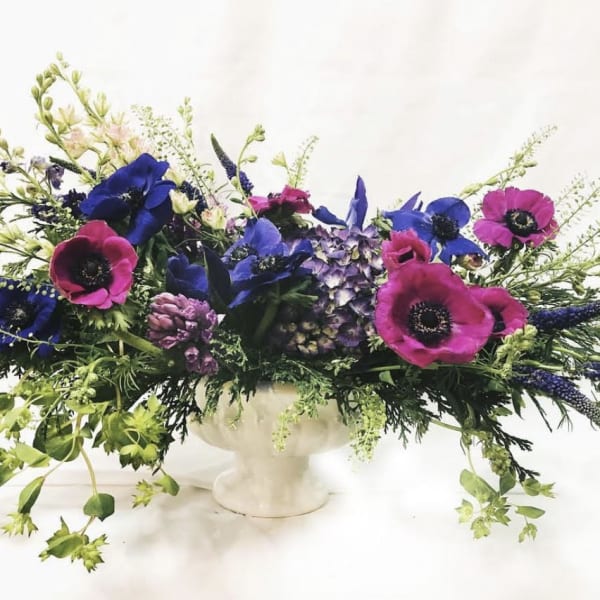 Long floral centerpiece with blue and magenta blooms in a white pedestal vase