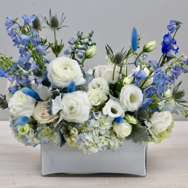 Low arrangement of white roses and ranunculus with blue delphinium in a gray ceramic vase