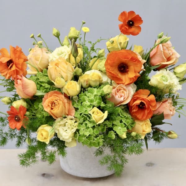 Low centerpiece of orange ranunculus, peach and yellow roses, and green hydrangeas in a white pot.