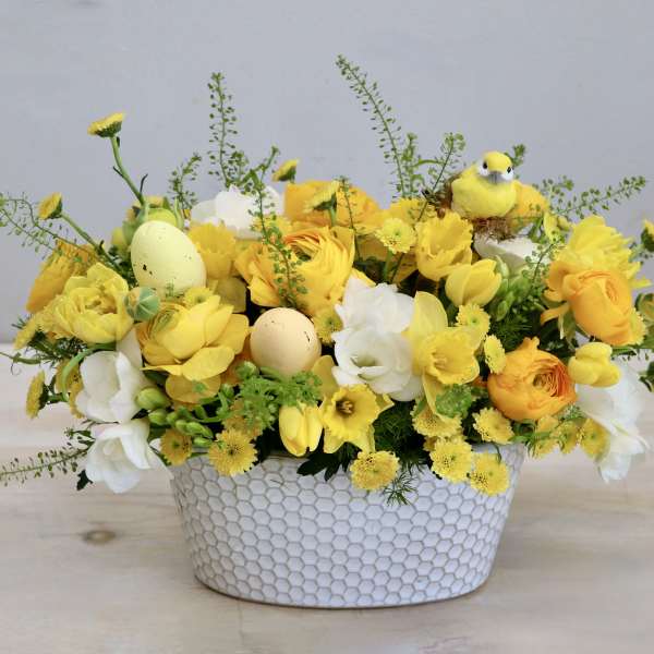 Low yellow and white flower arrangement with pastel eggs and a bird in a white textured ceramic container