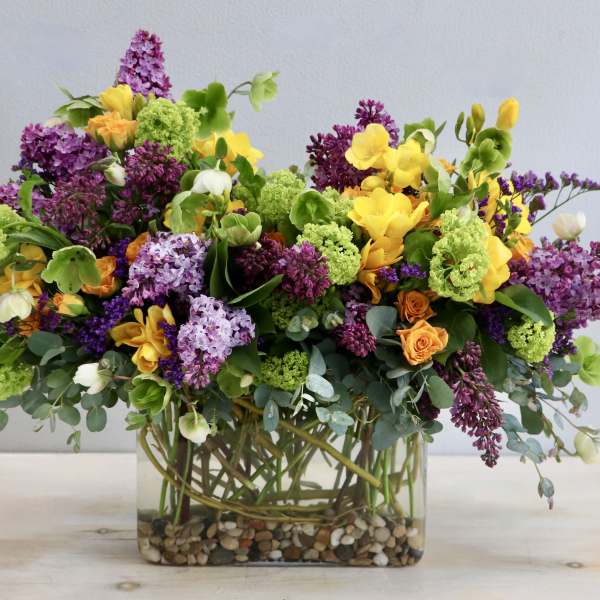 Lush mixed arrangement of purple, yellow, green, and orange flowers in a rectangular glass vase with pebbles.
