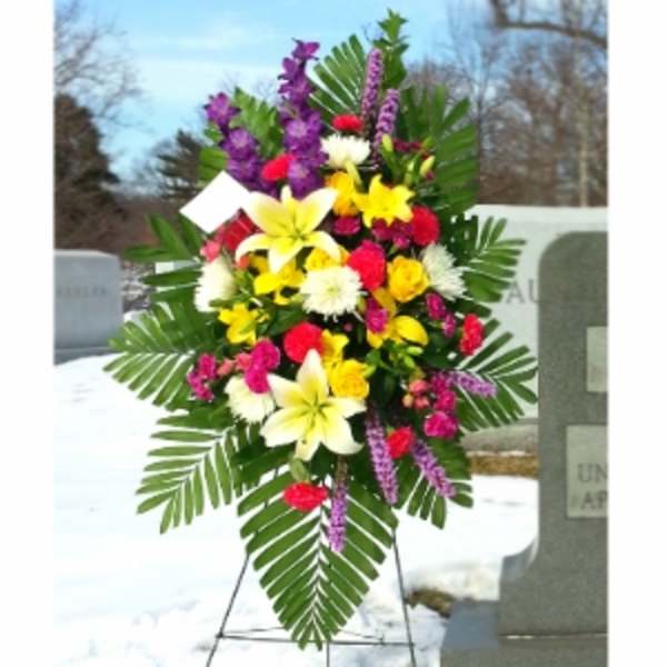 Standing spray of yellow lilies, bright pink flowers, and white mums on an easel in a cemetery