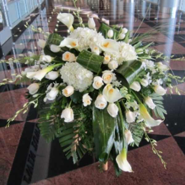 Large white arrangement of roses, calla lilies, and hydrangeas with bold green leaves on a stand.