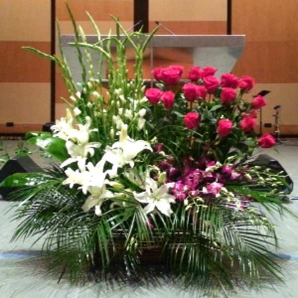 Large stage arrangement of white lilies, hot pink roses, and purple flowers with tall green spikes in a low container.