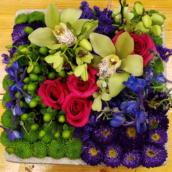 Square arrangement with pink roses, green orchids, purple mums and blue delphinium in a low container