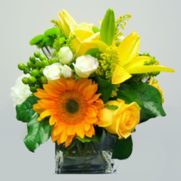 Bright arrangement with a sunflower, yellow lilies and roses, and white spray roses in a clear glass cube vase