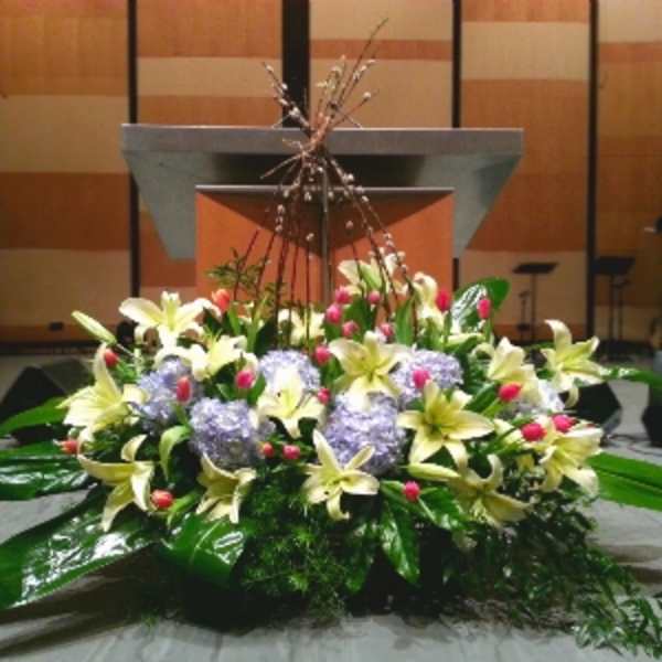 Low altar arrangement of yellow lilies, lavender hydrangeas, and pink buds with large glossy foliage.