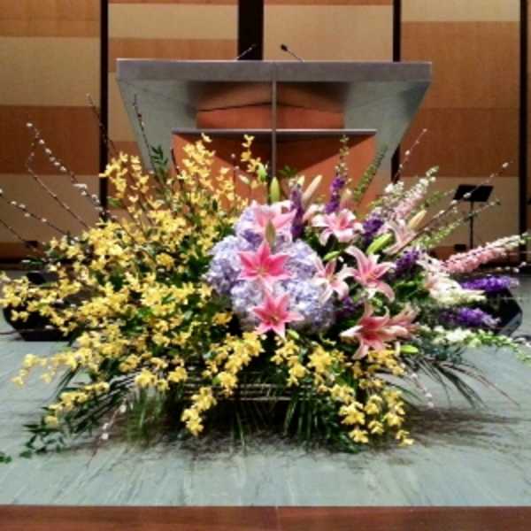 Large stage floral arrangement with pink lilies, lavender hydrangeas, yellow orchids, and tall accent stems