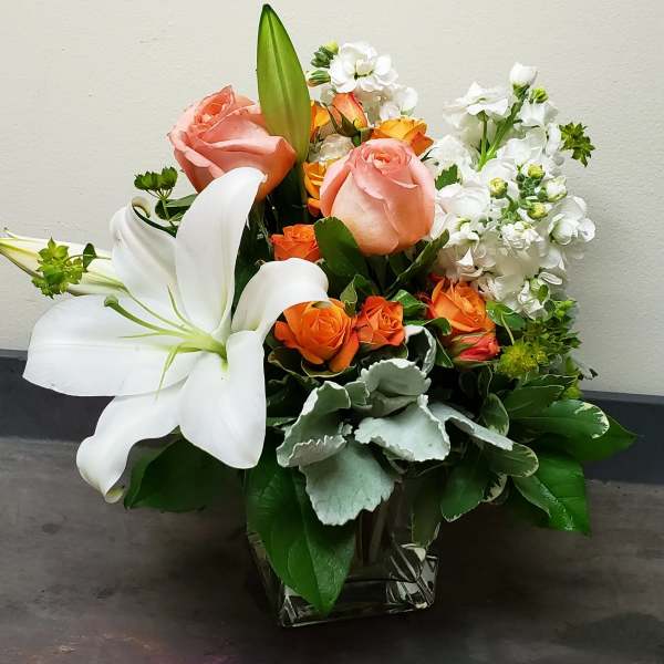 Compact arrangement of white lilies, peach roses, and orange spray roses in a square glass vase