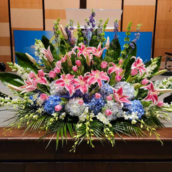 Large altar-style arrangement of pink lilies, blue hydrangeas, and white flowers in a fan shape