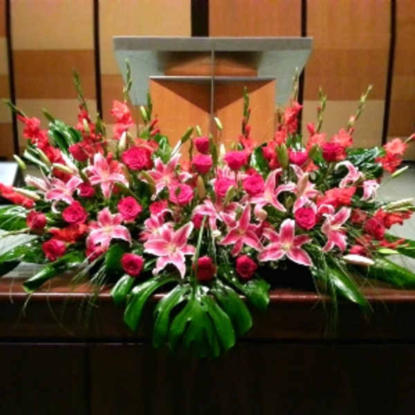 Large altar spray of pink lilies and red roses with bold tropical leaves on a wooden podium.