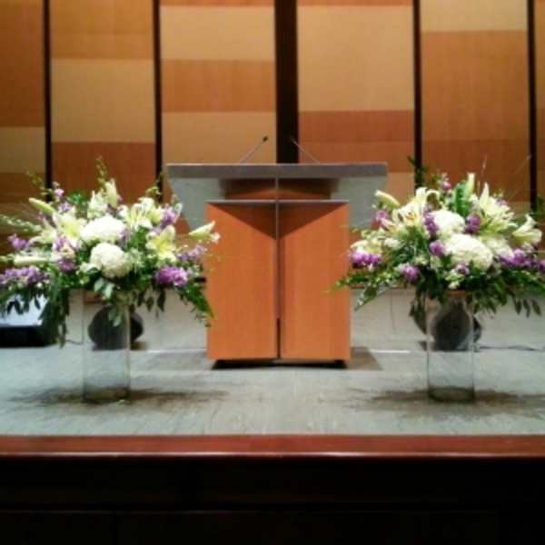 Two large white and purple floral arrangements in tall clear vases flanking a wooden podium