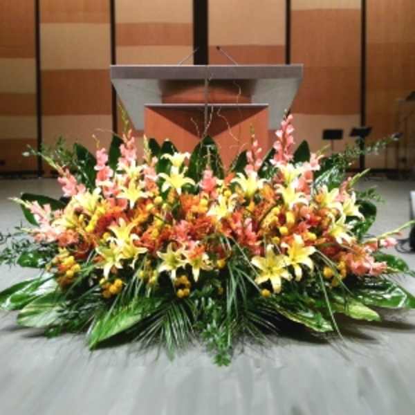 Low, wide arrangement of yellow lilies and peach flowers with large green foliage in front of a stage podium