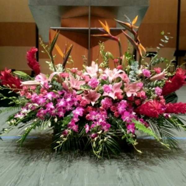 Large pink and red tropical altar spray with lilies and orchids arranged low and wide on a stage