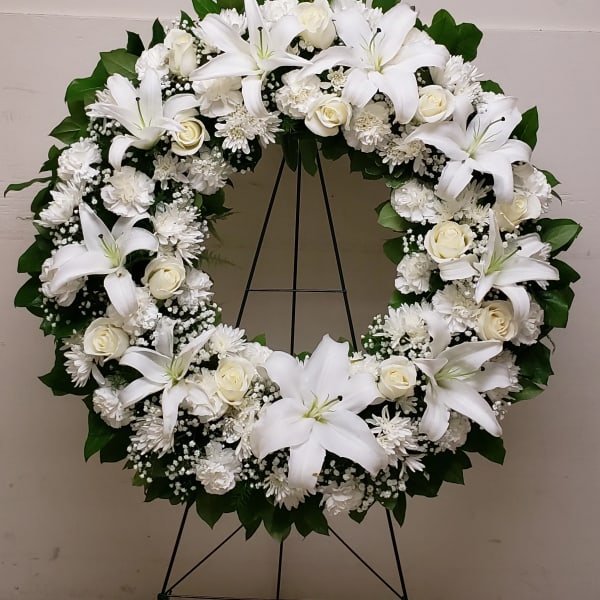 Large white floral wreath with lilies, roses, and mums displayed on a metal easel stand
