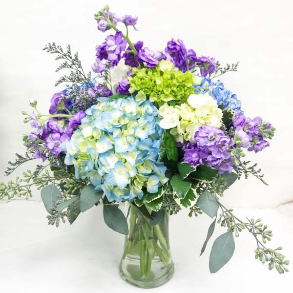 Arrangement of blue and white hydrangeas with purple stock in a clear glass vase