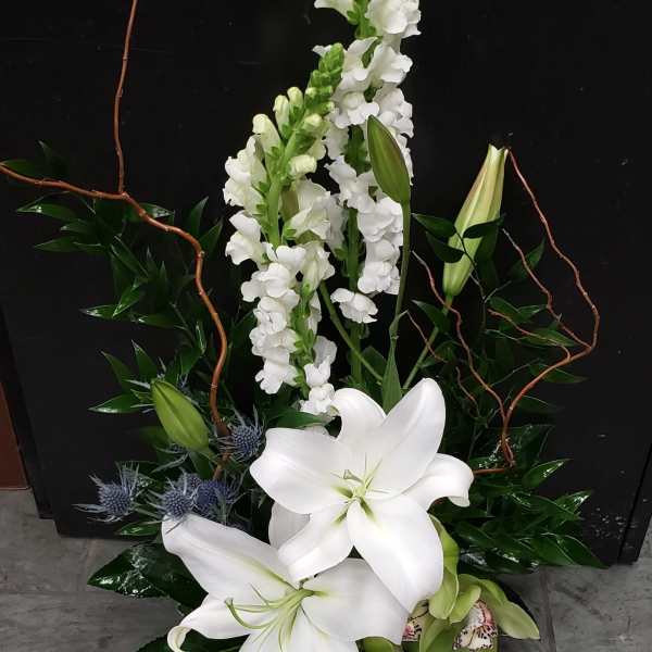 Tall arrangement of white lilies and snapdragons with blue thistle in a dark container
