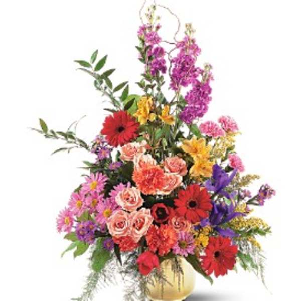 Tall mixed arrangement of pink roses, red gerbera daisies, carnations, and assorted bright flowers in a gold vase