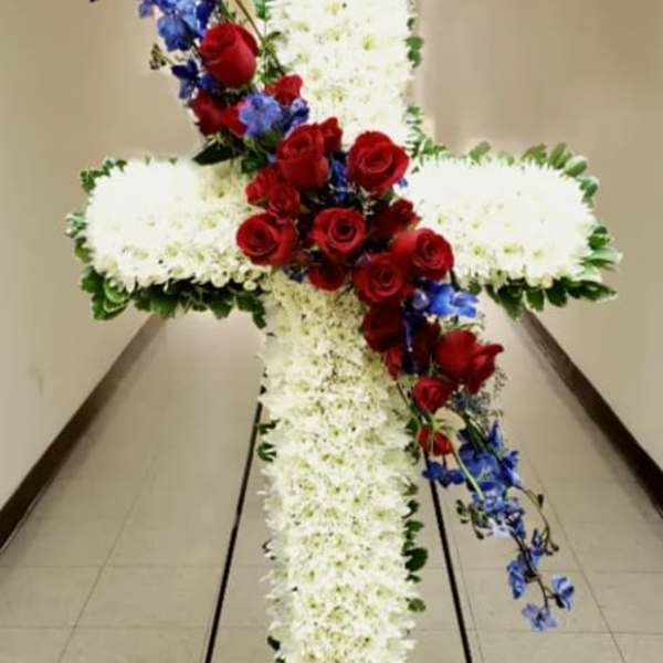 Tall white floral cross with red roses and blue flowers on a metal stand