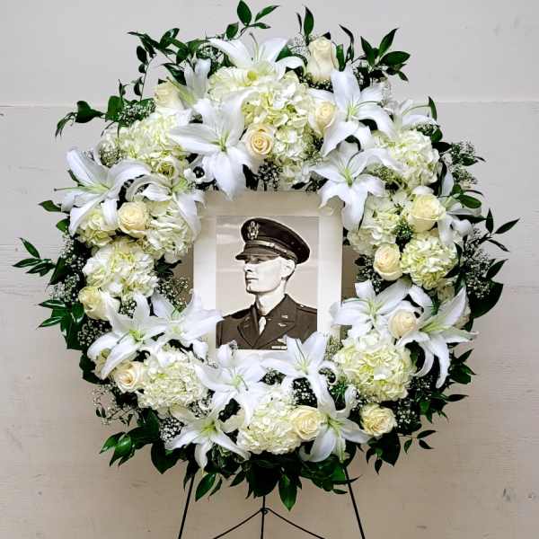 Large white floral wreath with lilies and roses on an easel framing a black-and-white portrait