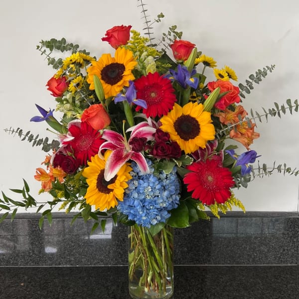 Mixed bouquet with sunflowers, roses, lilies, and blue hydrangea in a glass vase