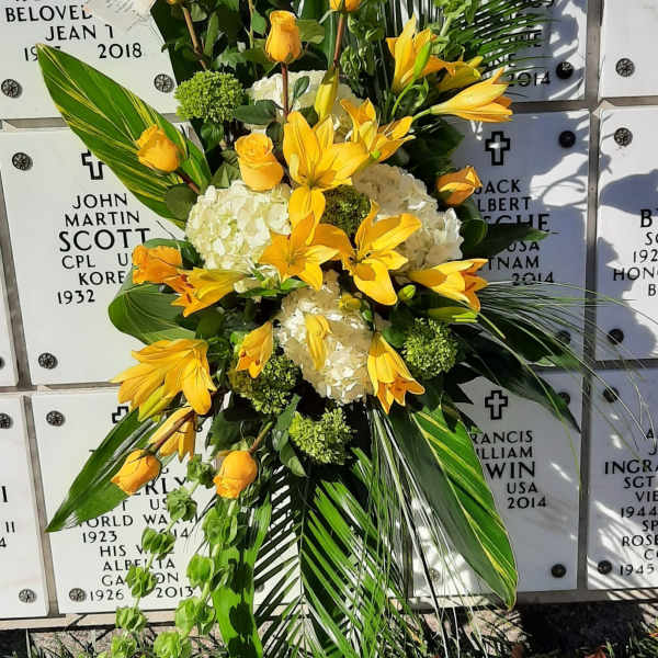 Tall spray of yellow lilies and roses with white hydrangeas on a stand in front of a marble memorial wall