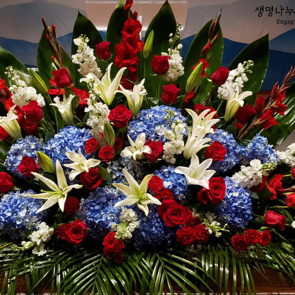 Large red, white, and blue floral spray with roses, lilies, hydrangeas, and bold green leaves.