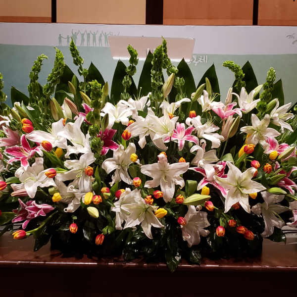 Large stage arrangement of white lilies, pink lilies, and yellow-orange tulips in a wide, low fan shape.