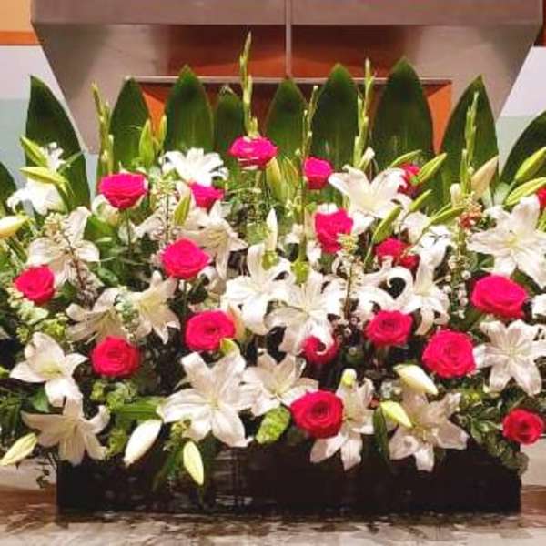 Large low arrangement of red roses and white lilies with bold green leaves on a stage.