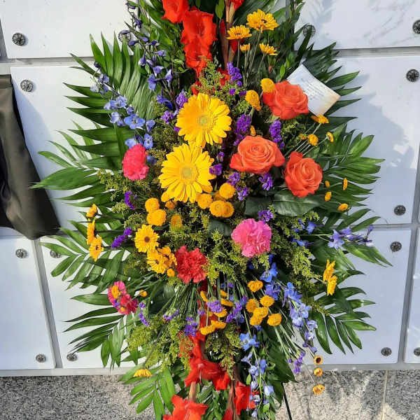 Tall standing spray of multicolored flowers including yellow gerbera, orange roses, and red gladiolus on an easel.