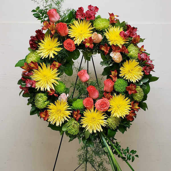 Standing wreath of yellow mums, pink roses, and red flowers on a green easel display