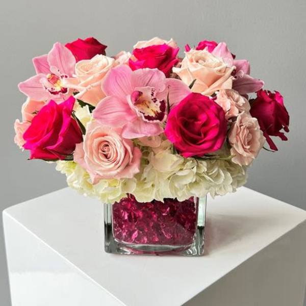 Compact arrangement of pink orchids, roses, and white hydrangeas in a clear cube vase