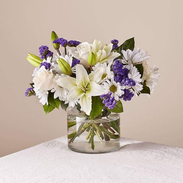 White lilies, daisies, and roses with purple accents in a clear glass vase.