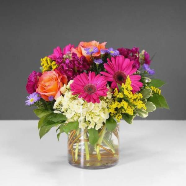 Bright mixed bouquet of pink gerbera daisies, orange roses, hydrangea, and filler flowers in a glass vase