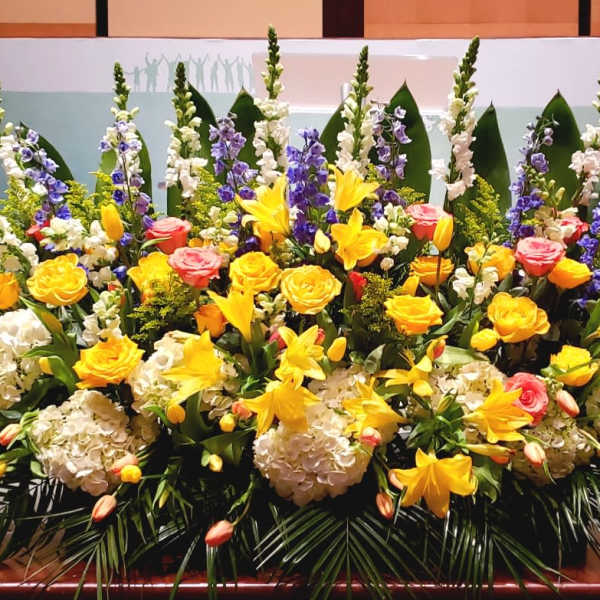 Large altar flower arrangement with yellow lilies and roses, white hydrangeas, pink roses, and blue delphinium.
