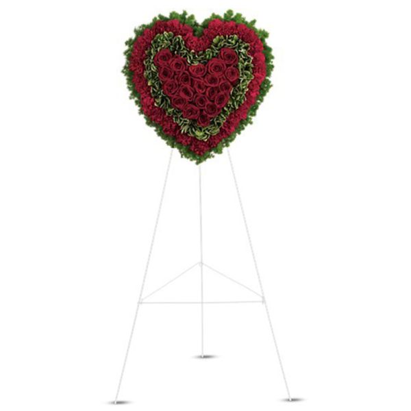 Heart-shaped red and green floral wreath displayed on a tall white easel stand.