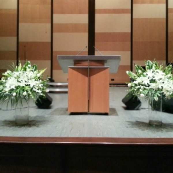 Two large white lily and rose arrangements in clear glass vases on a stage beside a podium